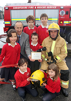 A posed group shot of 6 kids, headteacher, and firefighter in front of fire appliance