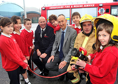A posed group shot of 6 kids, headteacher, and 2 firefighters in front of fire appliance