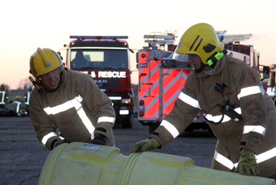 Two firefighters work with plastic drums in front of fire appliance