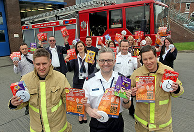 A group of fire service staff hold leaflets and smoke alarms towards the camera standing outside next to fire engine