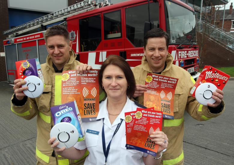 Three fire service personnel holding leaflets and smoke alarms in front of fire engine